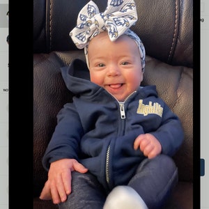 May include: A baby wearing a blue hoodie with a zipper and a white and blue bow with the word "Inghilte" on the hoodie. The baby is sitting on a brown leather chair.