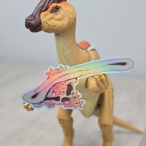 May include: A tan Parasaurolophus dinosaur toy holds a holographic sticker featuring a dinosaur skull with pink flowers. The dinosaur has a brown crest and orange accents. The sticker has a rainbow sheen.