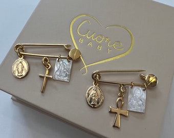 May include: Two gold safety pins with charms. The charms include a cross, an ankh, and two medallions with images of the Virgin Mary. The pins are in a box with the words "Cuore Baby" printed on it.