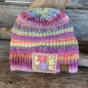 May include: A multi-colored knit beanie with a leather patch featuring three embroidered flowers. The patch has the text "and so is" and a small arrow. The beanie has a "CC" logo on the side.
