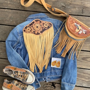 May include: A denim jacket with a brown leather fringe and floral design on the back. A brown leather crossbody bag with fringe and a floral design. A pair of brown leather earrings with a floral design. A pair of canvas shoes with a brown leather floral design. The shoes are on a wooden surface.