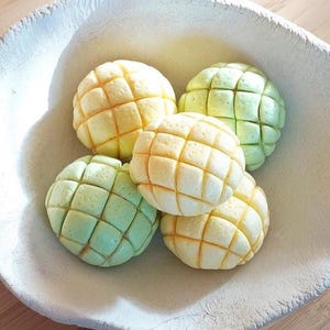 May include: A white, textured ceramic bowl holds five melon pan pastries. The pastries are a mix of yellow and green, with a grid-like pattern on top. The bowl sits on a wooden surface.
