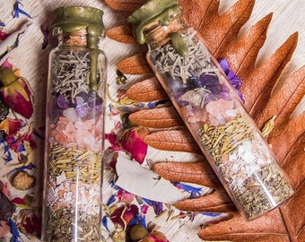 May include: Two small glass bottles with cork stoppers and gold lids filled with various herbs, crystals, and dried flowers. The bottles are sitting on a wooden surface with dried leaves and flowers.