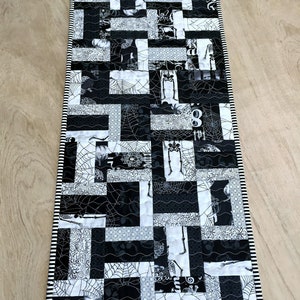 May include: A rectangular table runner featuring a patchwork design of black and white fabrics with Halloween-themed prints including spider webs, skeletons, and skulls. The runner is finished with a black and white striped fabric border.