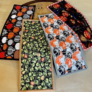 May include: Three rectangular fabric table runners featuring seasonal patterns. One displays orange, white, and black pumpkins on a black background. Another shows green ivy leaves and small pumpkins. The third features glowing jack-o'-lanterns.