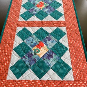 May include: A quilted table runner featuring a repeating pattern of teal and white diamonds, with floral fabric squares at the center. The border is a rust-colored fabric with a small white floral pattern, and a teal green edge.