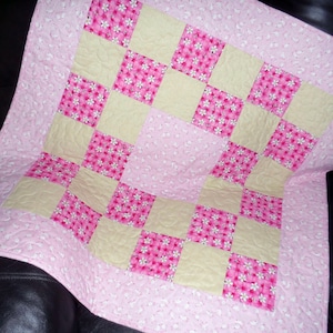 May include: A pink and cream-colored baby quilt with a checkerboard pattern. The quilt features alternating squares of pink fabric with white floral designs and cream-colored squares. The edges are finished with a pink floral patterned fabric.