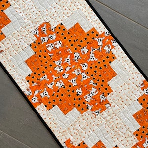 May include: A Halloween-themed table runner with a black border. The runner is made of orange fabric with a white polka dot pattern and a ghost print. The center of the runner is made of white fabric with a paisley pattern.