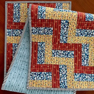 May include: A quilted table runner with a patchwork design. The runner features a combination of red, yellow, and blue floral patterns. The edges are finished with a black and white striped border, and the backing is a light blue fabric with a star pattern.
