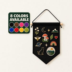 Op de afbeelding: Een zwarte stoffen wandbanner met een houten stok en ophangkoord, voorzien van diverse emaille pins waaronder bloemen, een maansikkel, bergen, een bij, paddenstoelen, een blad, een hart, een planeet, een regenboog, een kat en een ronde bergpatch.