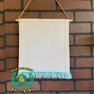 Puede incluir: Un tapiz de pared rectangular de lona blanquecina con flecos azul claro en la parte inferior. La lona está suspendida de un palito de madera y un cordón de hilo natural. El fondo es una pared de ladrillos.