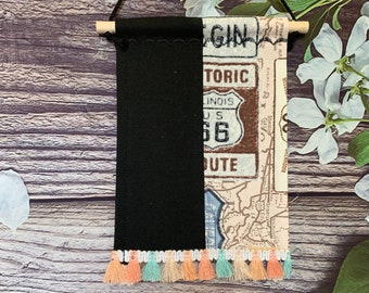 May include: A black and white fabric wall hanging with a vintage Route 66 design. The hanging has a black fabric panel on the left side and a white fabric panel on the right side with a vintage map and a Route 66 sign. The hanging has a fringe of peach, light blue, and white tassels.