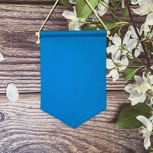 May include: A blank, turquoise blue pennant banner with a scalloped top edge, suspended by a natural twine cord and wooden dowel. The banner hangs against a rustic wooden background, with white flowers and green leaves in the corner.