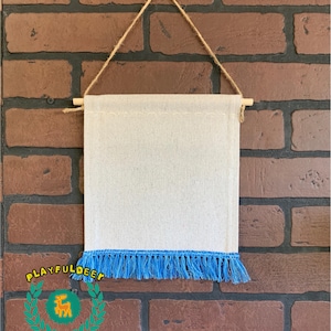 May include: A blank white canvas banner with a blue fringe hanging from a wooden dowel and twine. The banner is ready to be customized with your own design.