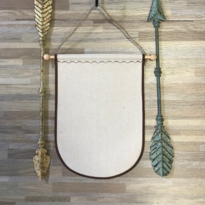 Könnte beinhalten: Rustikale Wanddekoration mit einem leeren Banner mit braunem Rand, flankiert von zwei dekorativen Metallpfeilen. Darüber hängt ein Holzblock mit Elefanten-Schnitzerei an einem Haken. Die Pfeile haben ein verwittertes, gealtertes Aussehen.