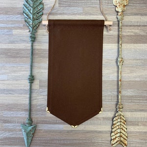 Op de afbeelding: Een bruine stoffen banner met gouden accenten hangt aan een touwtje tussen twee decoratieve metalen pijlen. De pijlen zijn geschilderd in lichtblauw en lichtbruin met roestkleurige accenten.