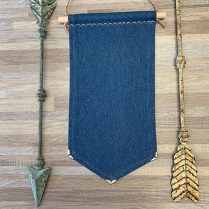 Pode incluir: Uma faixa de ganga com uma borda festonada pendura em uma haste de madeira com barbante. A faixa é ladeada por duas flechas de metal rústico, uma com um design de folha verde e a outra com uma ponta de flecha marrom.