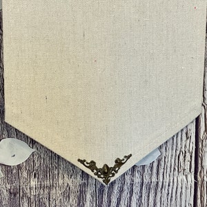 Op de afbeelding: Een beige canvasbanner met een decoratief metalen hoekstuk. De banner hangt aan een houten oppervlak.