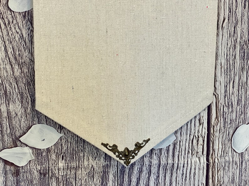 Op de afbeelding: Een beige canvasbanner met een decoratief metalen hoekstuk. De banner hangt aan een houten oppervlak.
