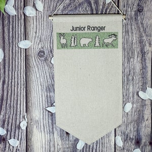 May include: A beige fabric banner with the text "Junior Ranger" printed in black. The banner has a green border with a deer, two trees, a bear, another tree, and a moose printed in black.