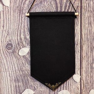 Könnte beinhalten: Schwarze Stoffbanner mit dekorativem goldenen Metallbesatz, das an einem Holzgrund hängt.
