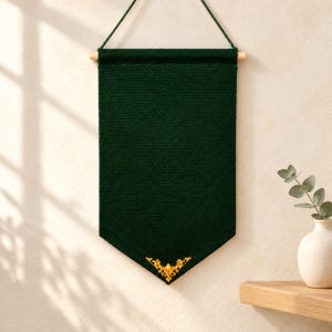 May include: A dark green fabric wall banner with a pointed bottom edge, suspended from a light-colored wooden dowel by a thin green cord. The banner features a decorative, ornate gold-colored metal embellishment centered at the bottom point.
