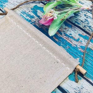 Op de afbeelding: Een blanco canvasbanner met een witte gestikte rand hangt aan een houten stok en touw. De banner is tegen een blauw en wit verweerd houten achtergrond. Een roze bloem met groene bladeren bevindt zich in de rechterbovenhoek van de afbeelding.