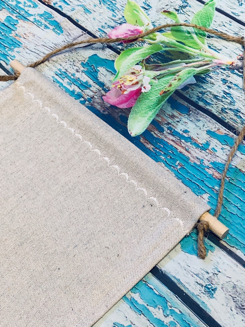 Op de afbeelding: Een blanco canvasbanner met een witte gestikte rand hangt aan een houten stok en touw. De banner is tegen een blauw en wit verweerd houten achtergrond. Een roze bloem met groene bladeren bevindt zich in de rechterbovenhoek van de afbeelding.