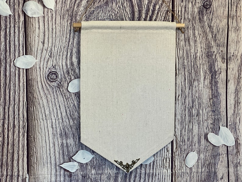 Op de afbeelding: Een blanco canvasbanner met een decoratieve metalen rand hangt aan een verweerde houten muur. De banner is gemaakt van natuurlijk linnen en heeft een rustieke, vintage uitstraling.