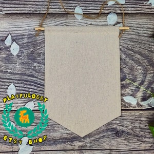 Può includere: Uno stendardo di tela bianca con una texture di lino naturale, appeso a un bastone di legno con uno spago. Lo stendardo è di colore beige chiaro e ha un bordo inferiore a punta. Lo stendardo è appeso su uno sfondo di legno rustico con petali di fiori bianchi sparsi intorno.