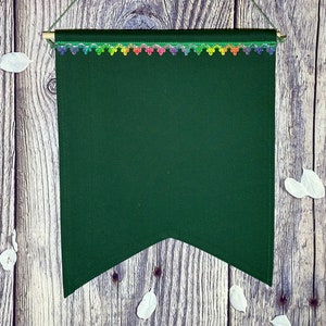 May include: A dark green fabric banner with a rainbow-colored trim hanging on a wooden wall. The banner is blank and has a pointed bottom.