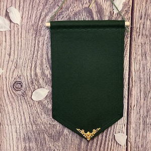 May include: A dark green fabric banner with a scalloped edge and a gold metal embellishment at the bottom. The banner is hanging from a wooden dowel rod.