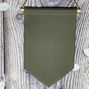 May include: A blank green canvas banner with a scalloped top edge and a pointed bottom edge. The banner is hanging from a wooden dowel rod with twine.