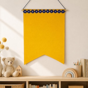 May include: A mustard yellow fabric wall banner with a pointed bottom edge, suspended from a wooden dowel by a black string. The top of the banner is decorated with a horizontal row of small, dark blue flower-shaped appliques.