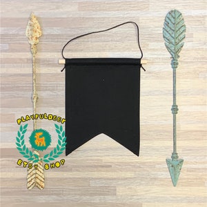 Op de afbeelding: Een zwarte stoffen banner met een puntige onderkant, hangend aan een houten lat met zwart touw. De banner is leeg en klaar om te worden gepersonaliseerd. Twee rustieke metalen pijlen met decoratieve punten bevinden zich aan weerszijden van de banner.