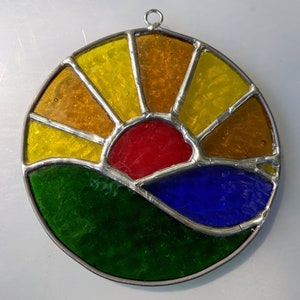 Könnte beinhalten: Ein Buntglas-Sonnenstrahl mit einer roten Sonne, blauem Wasser und grünem Gras. Der Sonnenstrahl ist in silberfarbenem Metall gefasst.