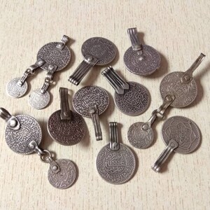 May include: A collection of silver-toned pendants featuring intricate Arabic calligraphy designs. Each pendant has a unique shape and size, with some featuring a hook or loop for hanging.