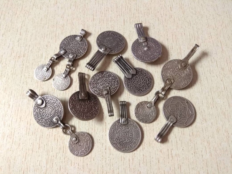 May include: A collection of silver-toned pendants featuring intricate Arabic calligraphy designs. Each pendant has a unique shape and size, with some featuring a hook or loop for hanging.