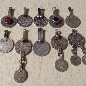 May include: A collection of 12 silver-toned pendants featuring intricate designs and Arabic script. Some pendants have red or black gemstones. The pendants are on a light wood surface.