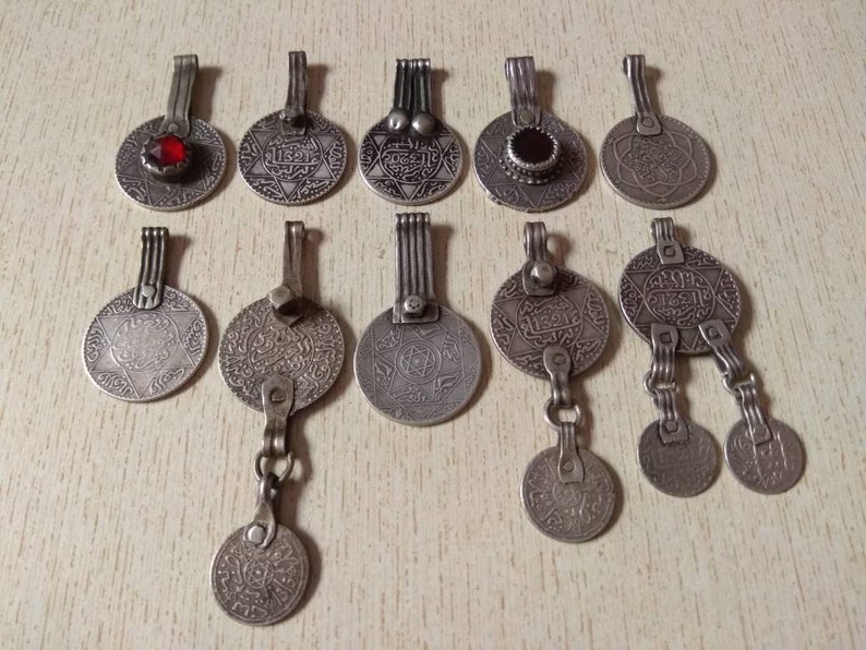 May include: A collection of 12 silver-toned pendants featuring intricate designs and Arabic script. Some pendants have red or black gemstones. The pendants are on a light wood surface.