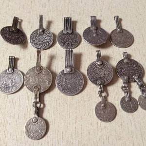 May include: A collection of twelve silver-toned pendants featuring intricate Arabic script and decorative designs. The pendants are circular and have a variety of styles, including some with multiple chains and charms.