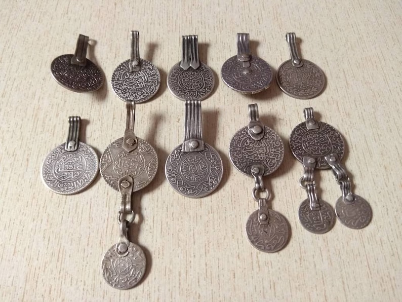 May include: A collection of twelve silver-toned pendants featuring intricate Arabic script and decorative designs. The pendants are circular and have a variety of styles, including some with multiple chains and charms.