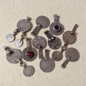 May include: A collection of silver-toned pendants featuring intricate Arabic script and red gemstones. The pendants come in a variety of designs, including circular, star-shaped, and geometric patterns.