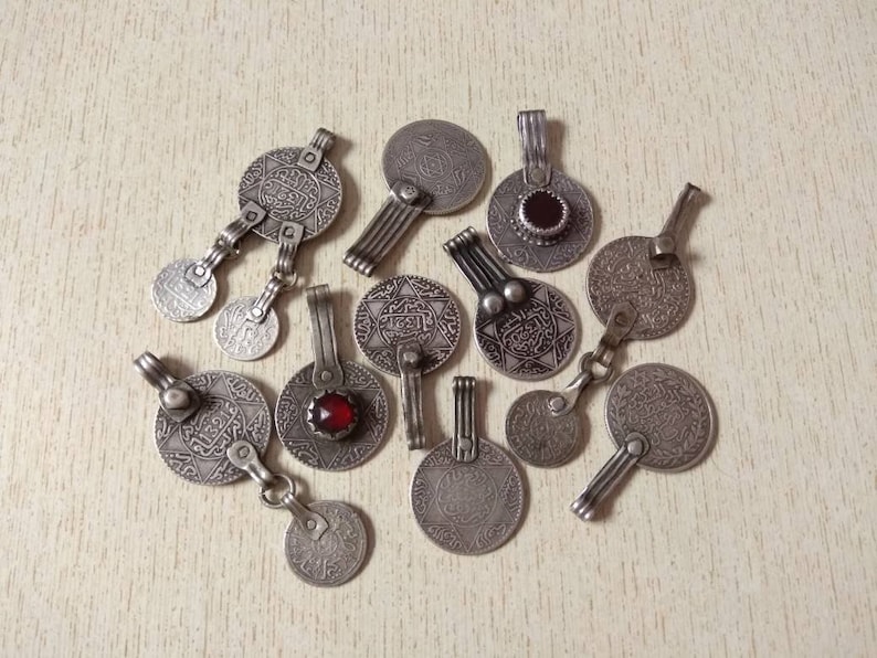 May include: A collection of silver-toned pendants featuring intricate Arabic script and red gemstones. The pendants come in a variety of designs, including circular, star-shaped, and geometric patterns.