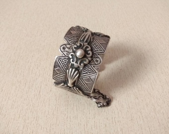 May include: A silver bracelet with intricate geometric designs and a small pearl accent. The bracelet has a chain attached to it.