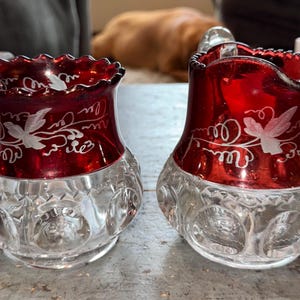 May include: Two antique ruby red glass vases with clear, patterned bases. The vases have a ruffled rim and etched white floral designs. The vases are likely decorative and vintage.