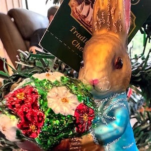 Puede incluir: Adorno navideño con un conejito sosteniendo un ramo de flores. El conejito está pintado en dorado, marrón y azul, con nariz y orejas rosas. Las flores son rojas y blancas. El adorno tiene una etiqueta que dice "Traditional Christmas".
