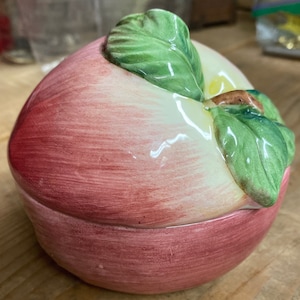 May include: A ceramic apple shaped trinket box with a green leaf and stem. The box is painted in shades of pink, white, and green.