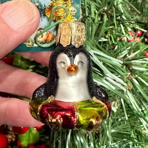 May include: A glass Christmas ornament shaped like a penguin. The penguin has a black body with glitter, a white belly, and a gold-colored crown. The ornament has red and green accents and a gold-colored metal hanger.