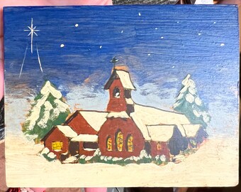 Puede incluir: Una tarjeta de Navidad de madera pintada a mano que representa una iglesia roja con un campanario y un techo cubierto de nieve. La iglesia está rodeada de dos árboles verdes con nieve blanca en sus ramas. El fondo es un cielo azul con estrellas blancas.